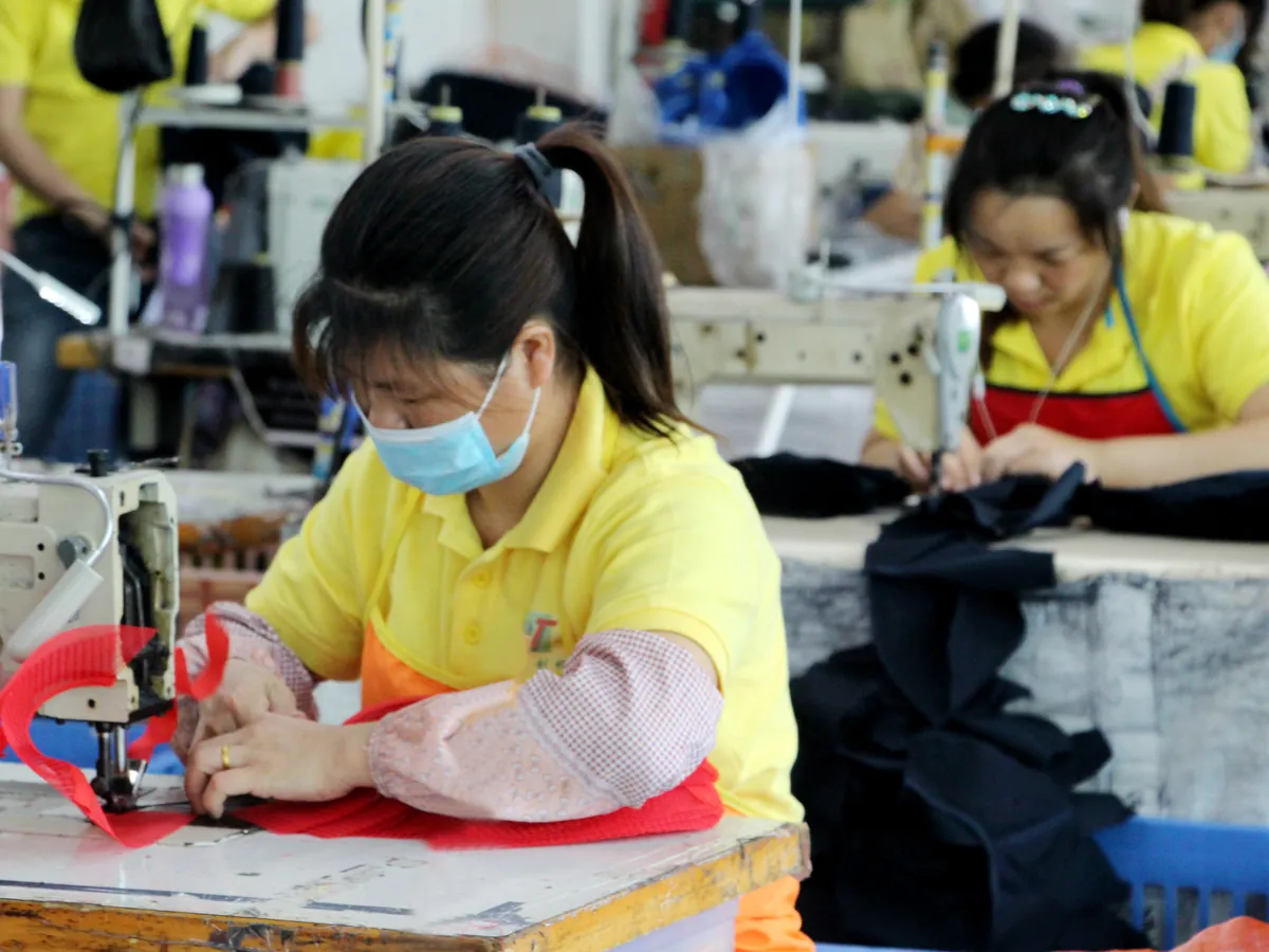Worker guiding red mesh fabric at a sewing machine, with another workstation in the background.