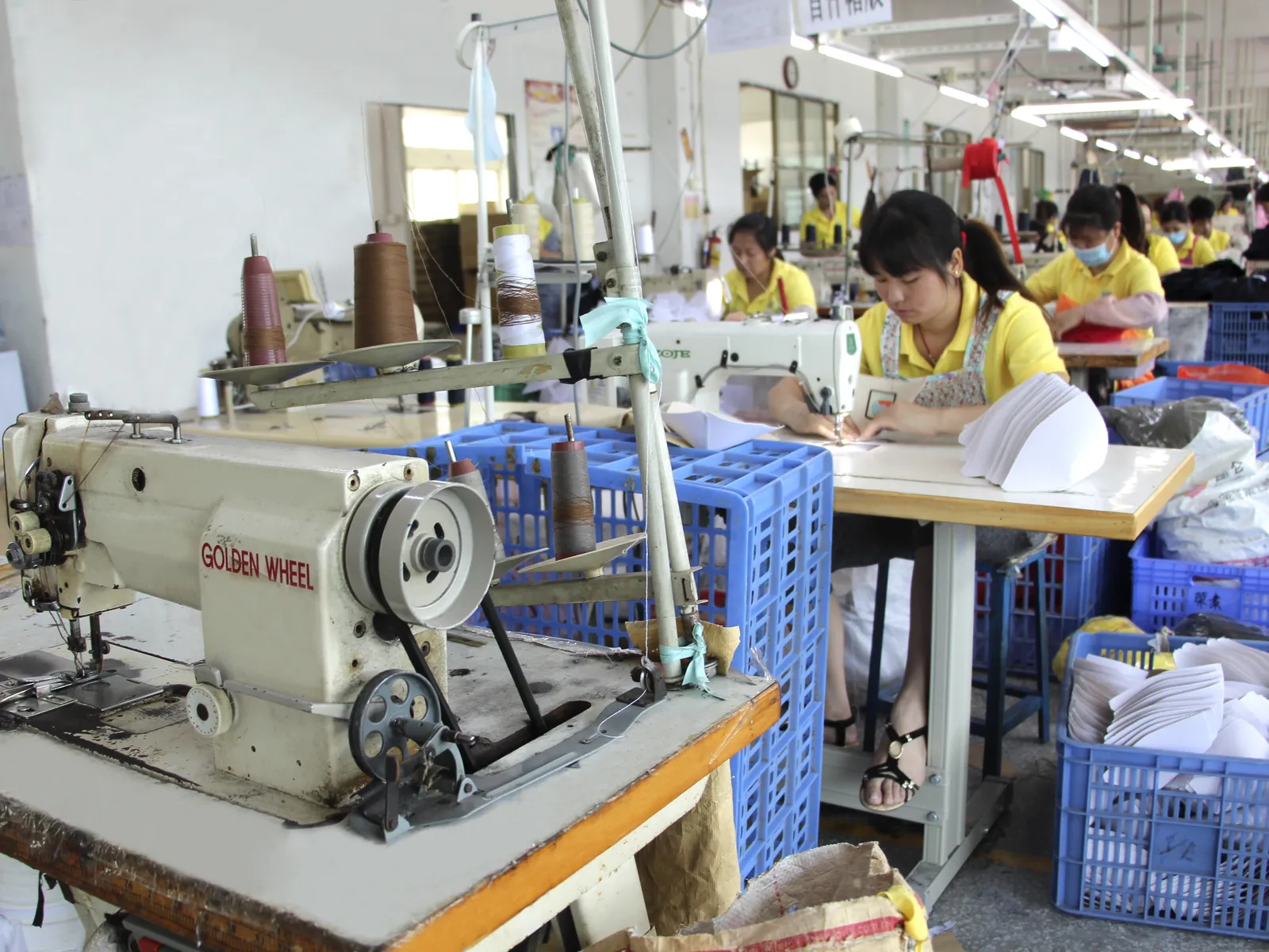 Workers sewing fabric components beside stacked white cap panels.